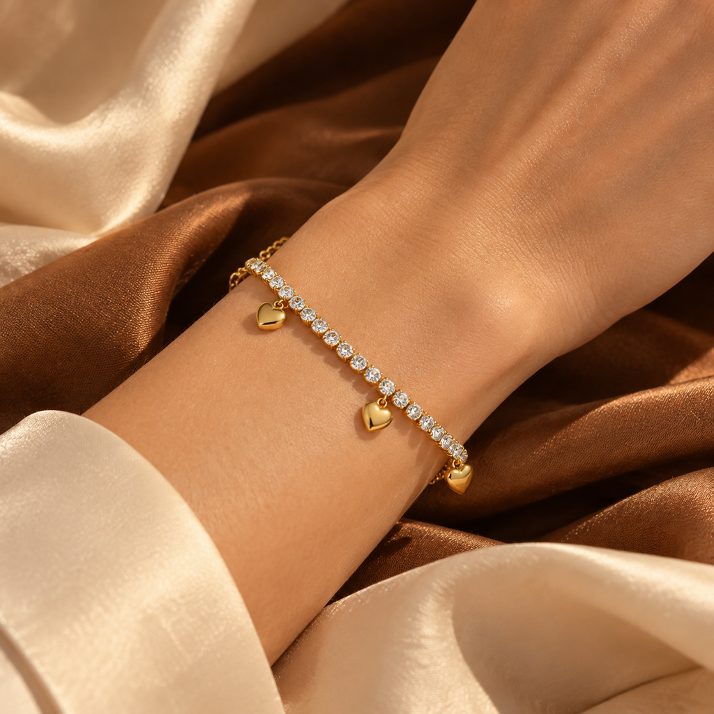 Model wearing the gold heart charm rhinestone tennis bracelet resting against an elegant bronze satin fabric.
