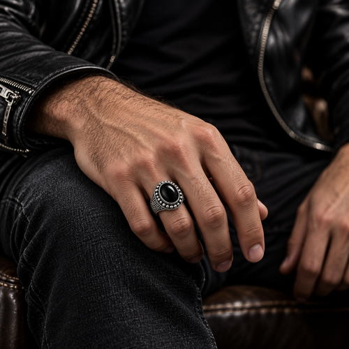 A close-up of a man's hand wearing a silver-toned ring with a large oval black stone, set against a dark leather jacket and jeans background.
