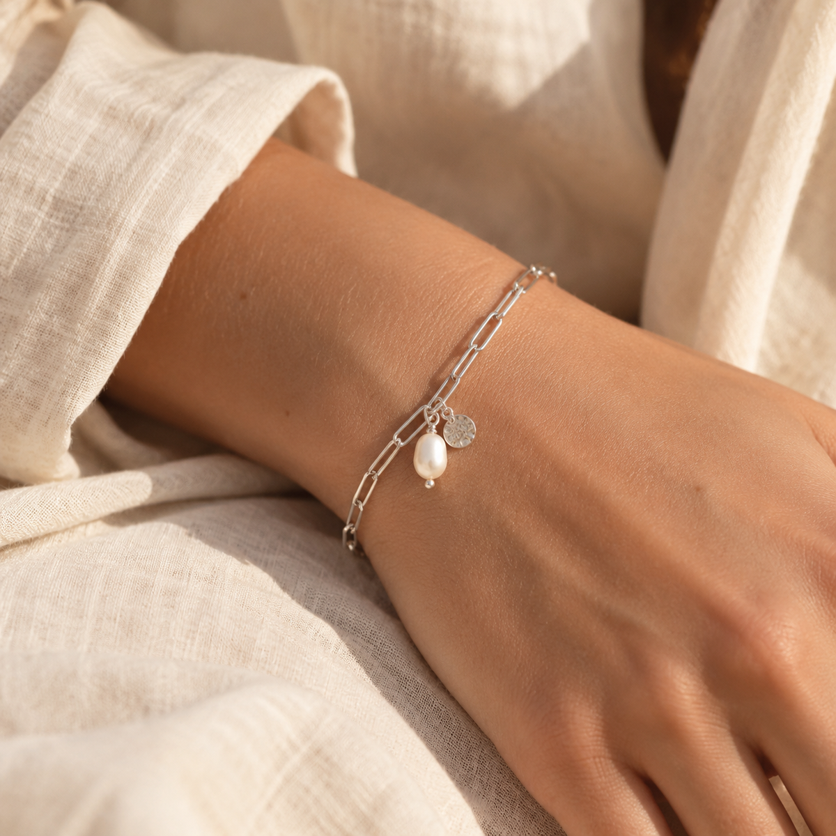 Close-up of woman wearing silver paperclip chain bracelet with dangling baroque pearl and hammered coin charm
