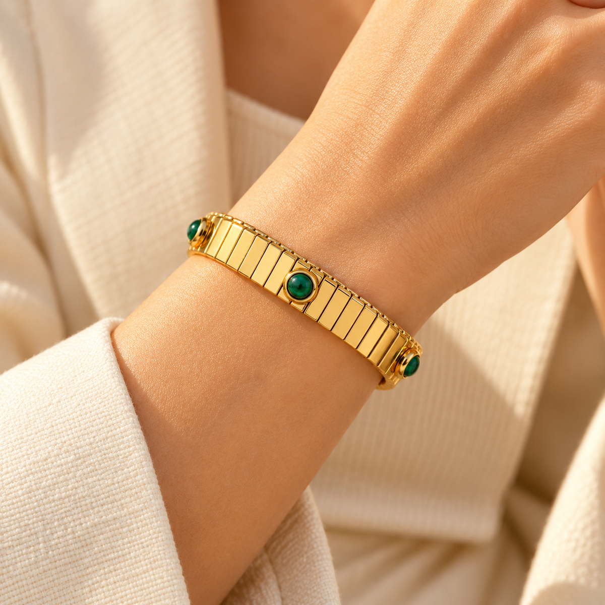 Close-up of model wrist wearing expandable gold watchband style stretch bracelet with green stone bezels and cream blazer
