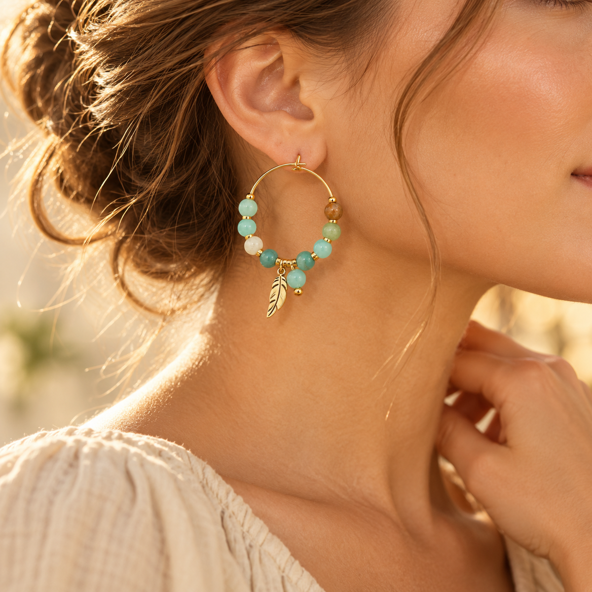 Close-up profile of model showcasing green natural stone beaded gold hoop earring with feather charm paired with cream knit top
