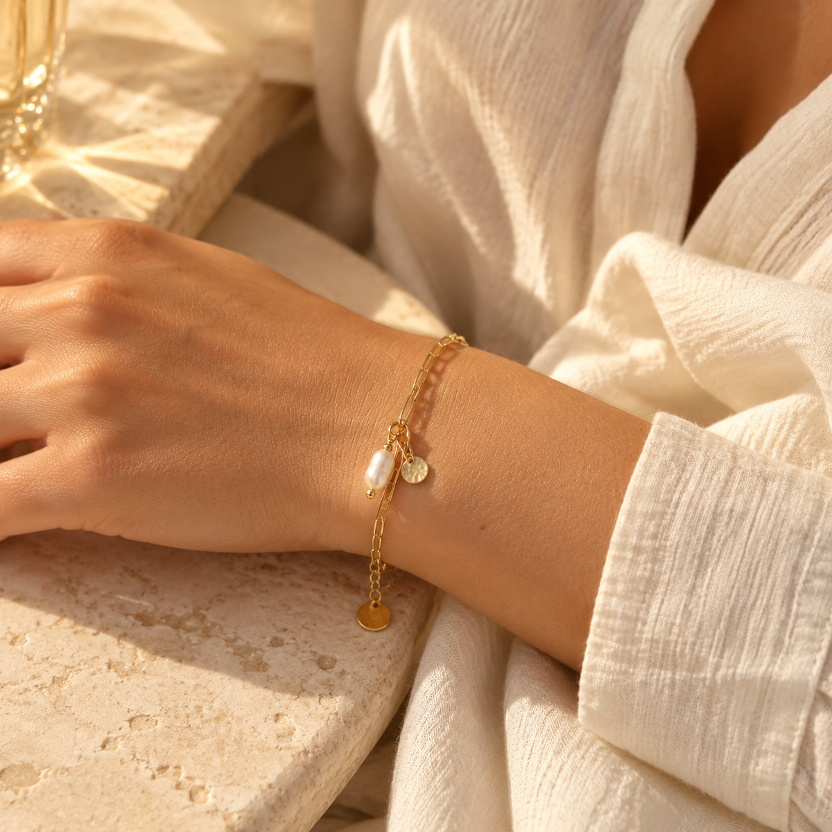 Lifestyle shot of woman wearing 18K gold plated paperclip chain bracelet with pearl charm resting on table
