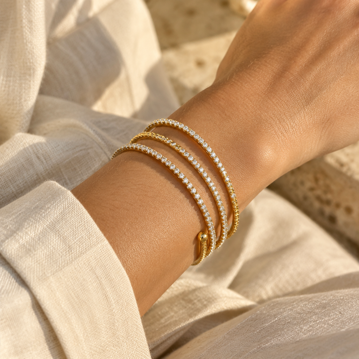 Close-up of a woman's wrist wearing the triple-layer gold spiral CZ tennis wrap bangle bracelet paired with an ivory linen shirt.
