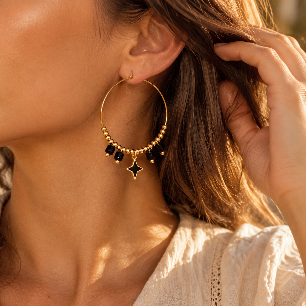 Side profile of woman wearing celestial gold hoop earrings with black beads and dangling black enamel starburst charm in sunlight
