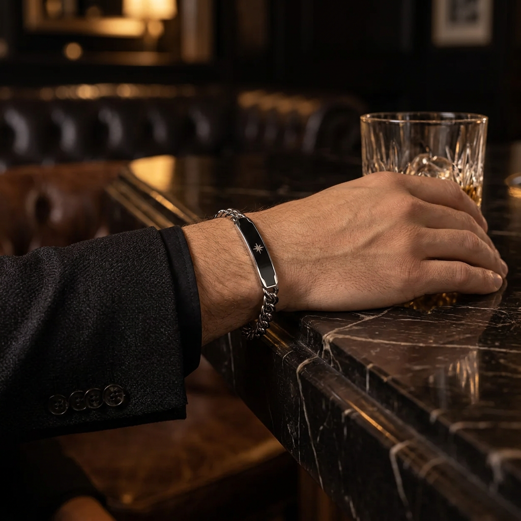 Close-up wrist view of men's black enamel Cuban ID bracelet with whiskey glass on marble bar
