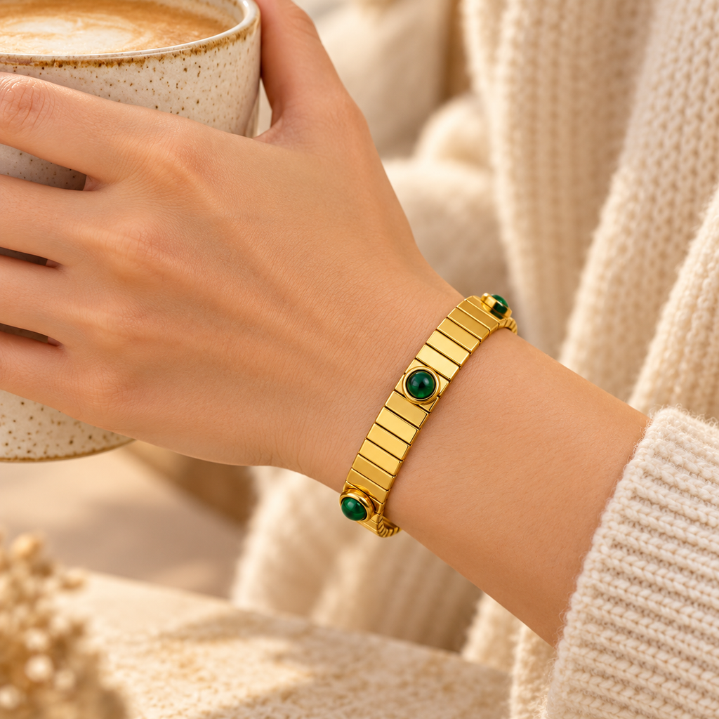 Side view of woman holding coffee cup displaying flexible gold ribbed bangle bracelet with emerald green accents
