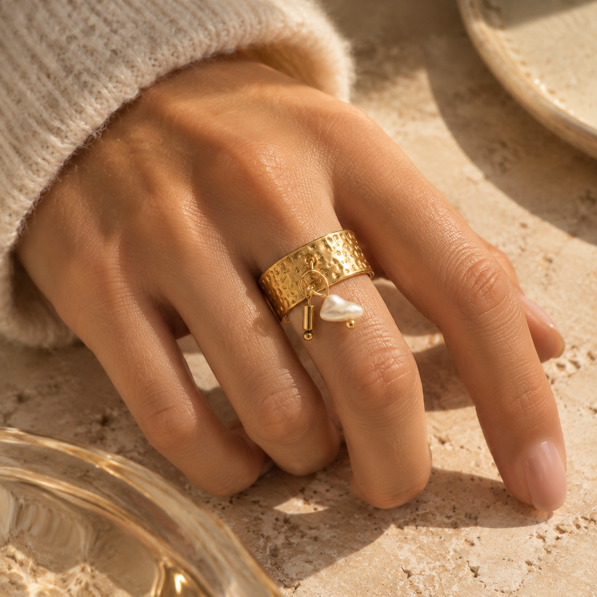 Aesthetic view of woman's hand wearing 18K gold plated wide band pearl charm ring on stone table
